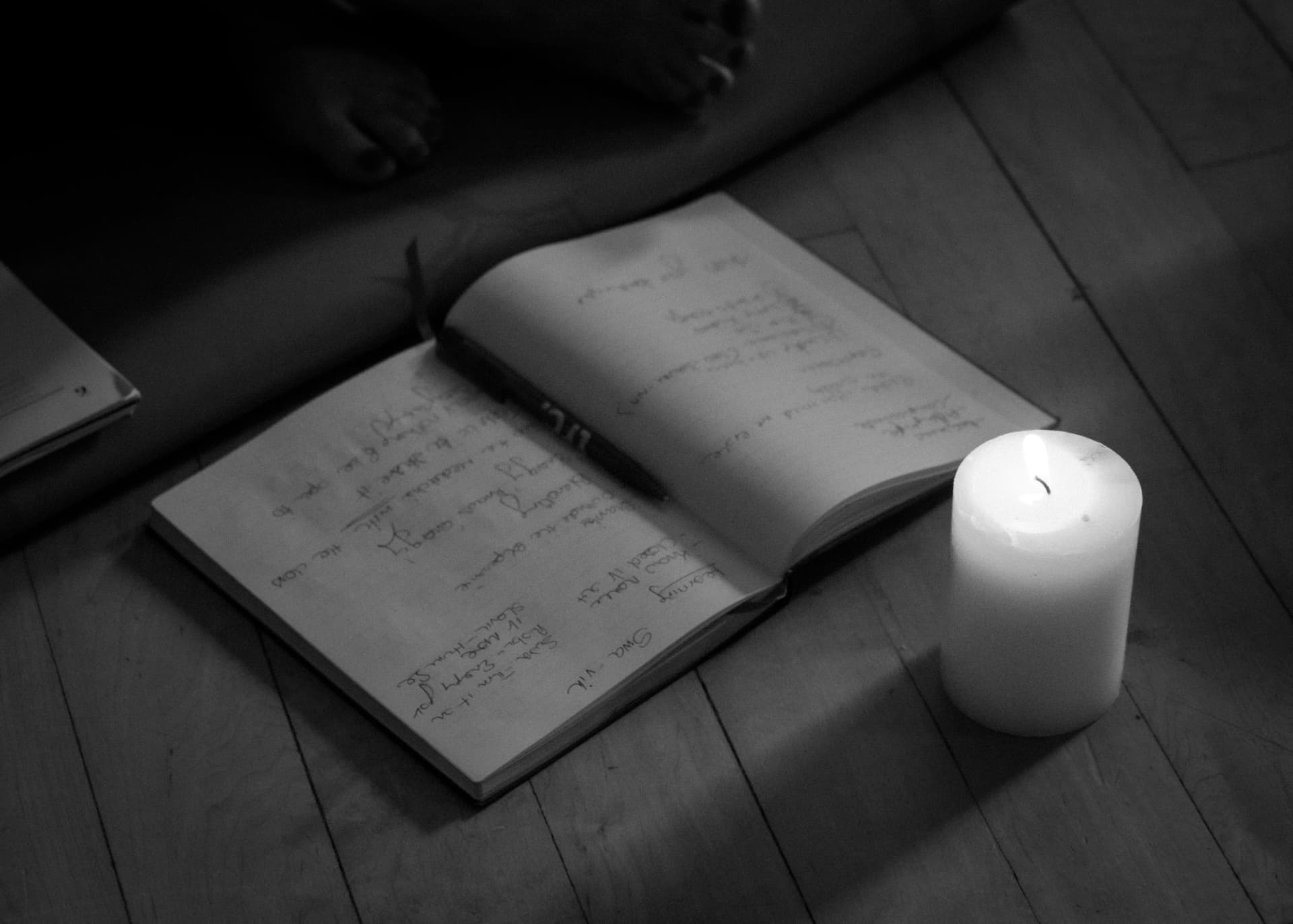An open handwritten journal, a pen and a lit white pillar candle on a wooden floor